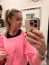 A woman taking a mirror selfie wearing a bright pink off-the-shoulder sweatshirt and a black undershirt. She is holding a smartphone with a beige case, and a framed picture is visible on the white wall behind her.