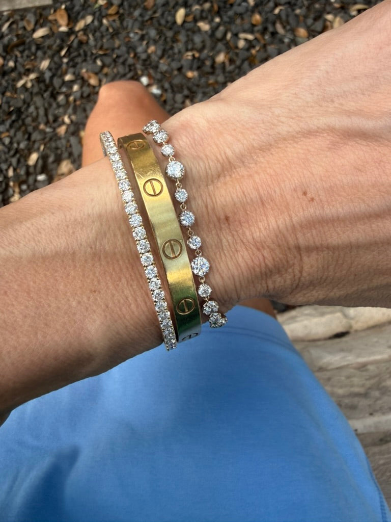 A close-up of a person's wrist wearing two bracelets: one gold-tone bracelet with screw details and one silver-tone pavé bracelet, with blue clothing and gravel in the background.