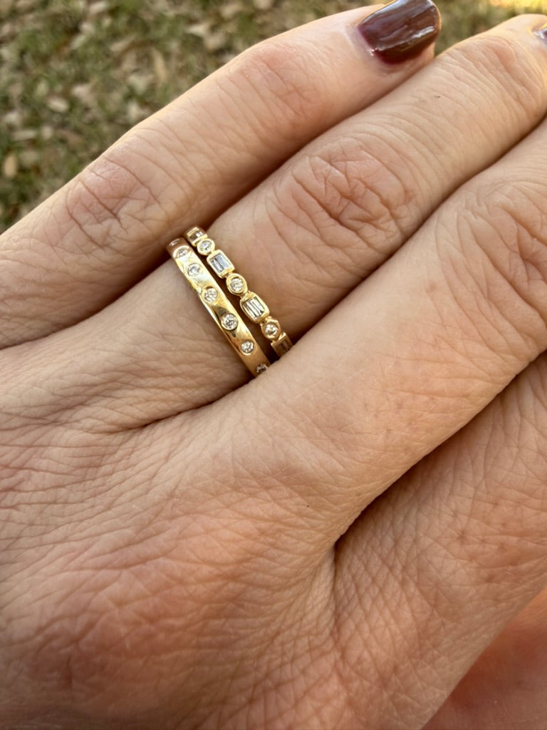 A close-up of a person's hand wearing two gold rings with small clear stones, photographed outdoors with blurred grass in the background.