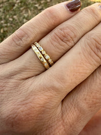 A close-up of a person's hand wearing two gold rings with small clear stones, photographed outdoors with blurred grass in the background.