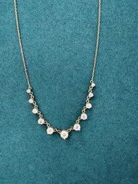 A delicate gold chain necklace featuring a V-shaped arrangement of small clear round-cut gemstones on a textured teal background.