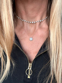 A close-up of a person wearing layered silver necklaces, including a choker with small clear stones and a delicate necklace with a flower-shaped pendant, paired with a black zip-up top.