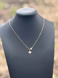 A delicate gold necklace with two small starburst pendants displayed on a matte black jewelry bust against a softly blurred outdoor background.