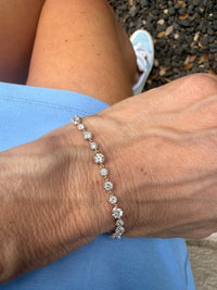 A close-up of a person's wrist wearing a delicate silver tennis bracelet with round-cut clear stones, set against blue clothing and dark gravel.
