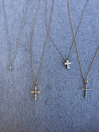Four gold-tone cross pendant necklaces with varying diamond-studded cross pendants, laid out on a textured gray fabric background.
