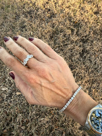 A close-up of a person's hand wearing a silver ring and a matching silver tennis bracelet, set against a background of dry grass outdoors.