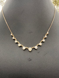 A delicate gold chain necklace with small heart-shaped gold pendants, each set with a clear round gemstone, displayed on a textured black jewelry bust.