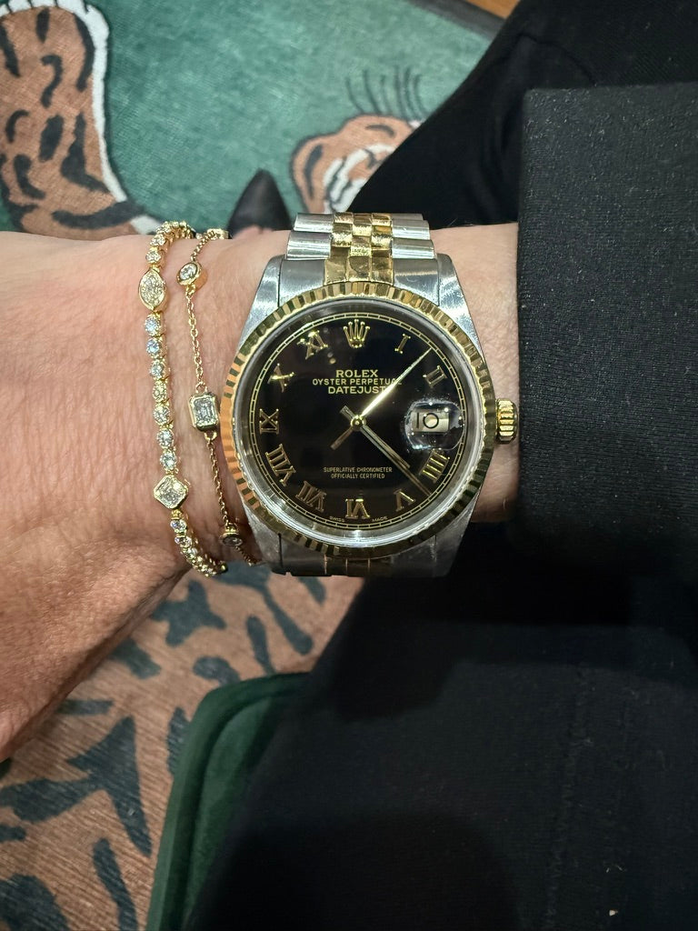A Rolex Datejust watch with a two-tone silver and gold stainless steel case and a black dial, worn on a person's wrist alongside gold bracelets.