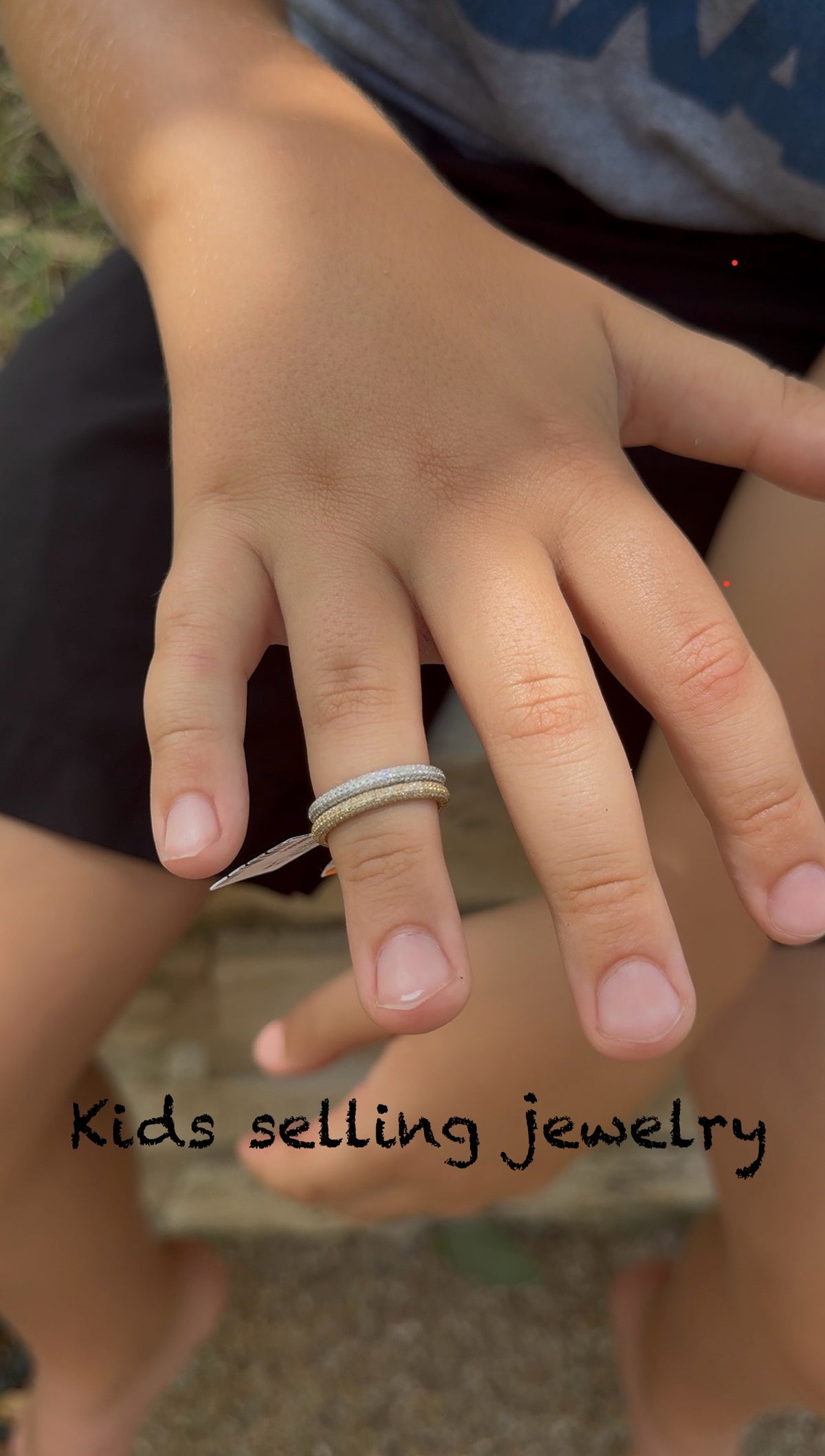 Close-up of a child's hand wearing a white gold ring with text 'Kids selling jewelry'.