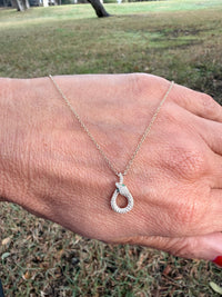A gold chain necklace with a small, diamond-encrusted teardrop-shaped pendant, displayed on a person's hand with an outdoor grassy background.
