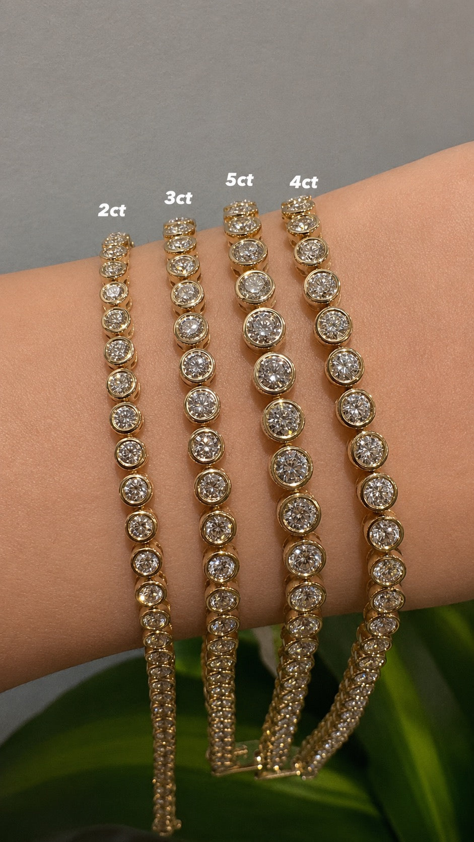 Four gold-toned tennis bracelets with different diamond counts (2ct, 3ct, 5ct, 4ct) displayed on a person's wrist against a gray background with green leaves at the bottom.