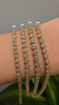 Four gold-toned tennis bracelets with different diamond counts (2ct, 3ct, 5ct, 4ct) displayed on a person's wrist against a gray background with green leaves at the bottom.