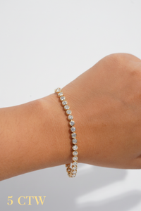 A bezel set  gold tennis bracelet with small diamonds, shown worn on a person's wrist against a clean white background.