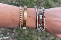 A close-up of a person's wrist wearing multiple bracelets outdoors. The bracelets include a gold bangle with screw details, a silver tennis bracelet, and several stacked rose gold and silver bangles, including one with the text 'I'S WILL'.