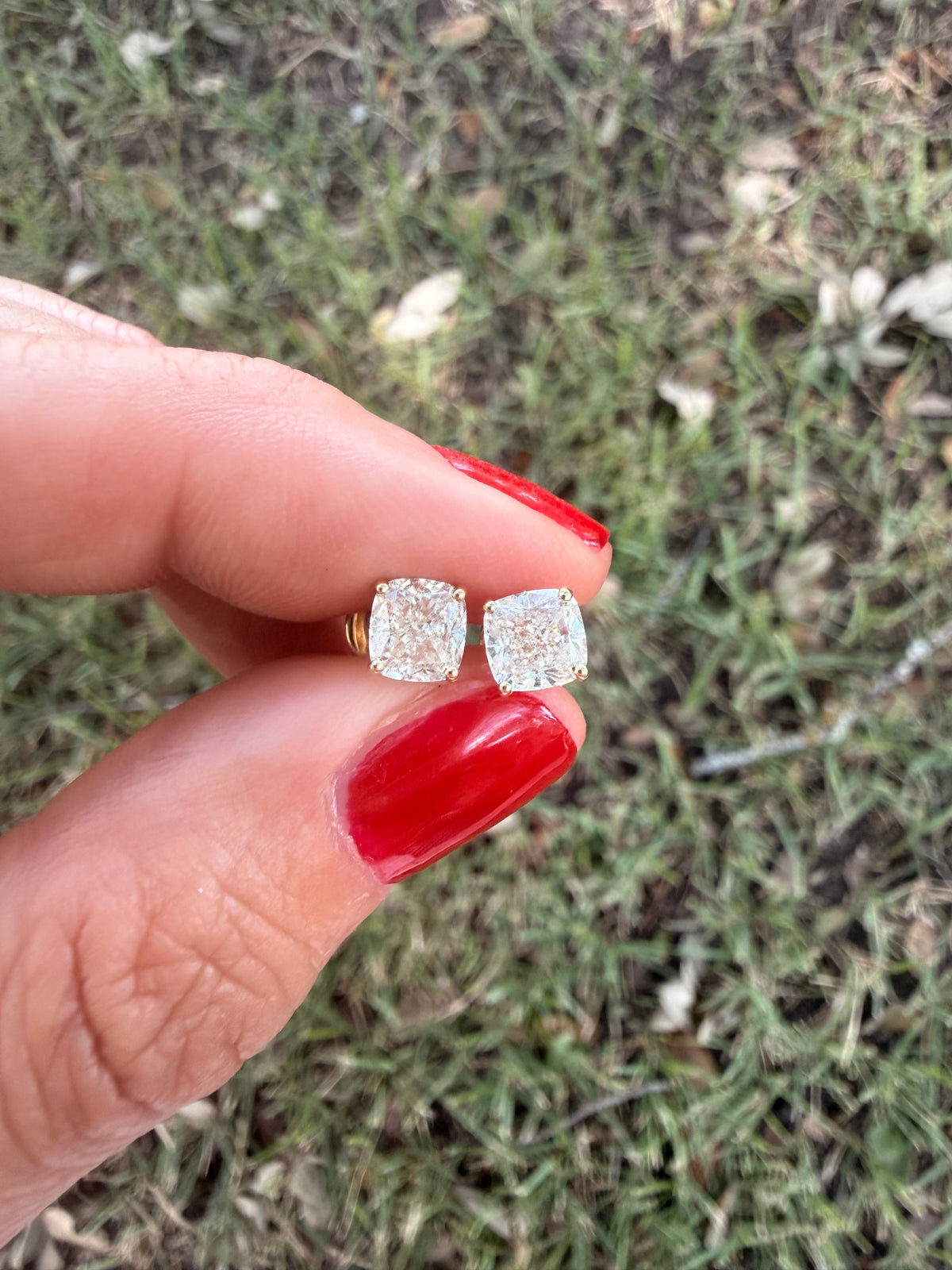 The Karat Concierge cushion cut diamond stud earrings in 18K yellow gold held between fingers with red nail polish.