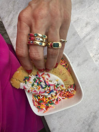 The Karat Concierge 14KT yellow gold donut rings stacked with colorful oval gemstones held over a sprinkle cookie.