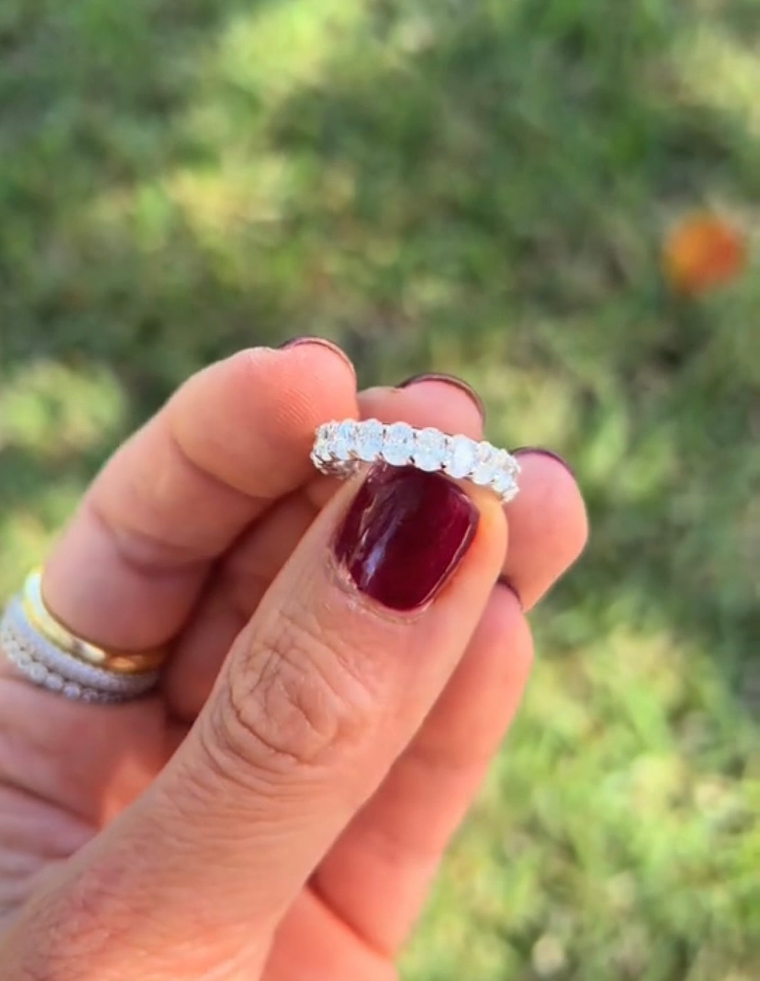 The Karat Concierge oval lab grown diamond eternity band held in hand against a blurred green outdoor background.